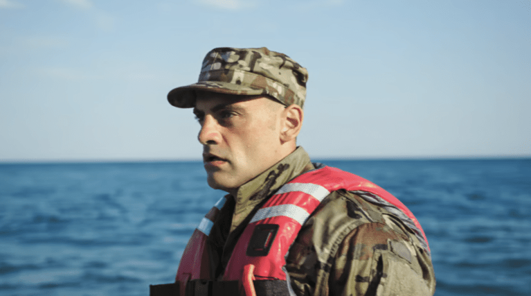 A man in camouflage uniform and a red life jacket stands by the sea, looking focused. The sky is clear and the water is calm in the background.