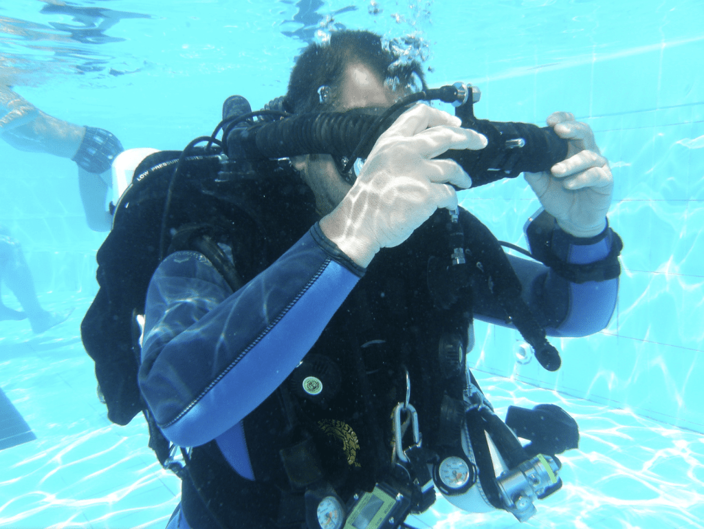 Screenshot 2026-03-30 at 165459 - N9BO℠ | Global Underwater Services Ltd A scuba diver in a wetsuit and gloves adjusts their mask whilst underwater in a pool, with diving equipment and gauges visible on their chest.