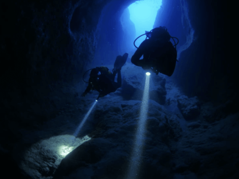 Two scuba divers explore an underwater cave, each shining a torch onto the rocky floor beneath them as blue light filters from the cave entrance above.
