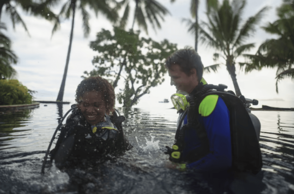 Screenshot 2026-03-31 at 184543 - N9BO℠ | Global Underwater Services Ltd Two people in scuba gear stand in shallow water, smiling and splashing each other, with palm trees and tropical foliage in the background.