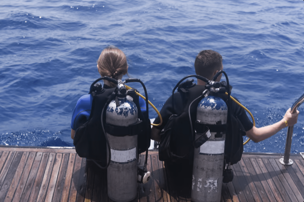 Screenshot 2026-03-31 at 184607 - N9BO℠ | Global Underwater Services Ltd Two people wearing scuba diving gear sit side by side on a wooden deck at the edge of a boat, facing the blue sea, preparing to enter the water.