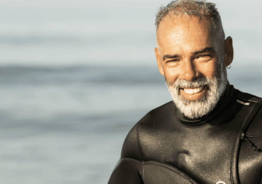 Screenshot 2026-03-31 at 184642 - N9BO℠ | Global Underwater Services Ltd A smiling man with grey hair and a beard wears a black wetsuit, standing by the sea with calm water in the background.