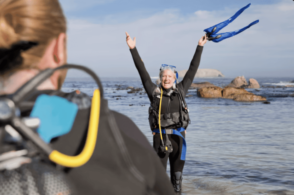 A person in scuba gear stands in shallow water with arms raised, holding flippers, smiling at another diver in the foreground. Rocky shoreline and sea are in the background under a blue sky.