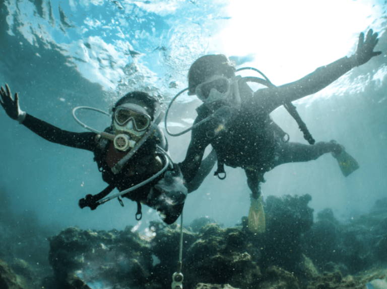 Two scuba divers wearing masks and wetsuits swim underwater over a rocky seabed, with sunlight filtering through the water above them. Both have their arms outstretched and appear to be enjoying the dive.