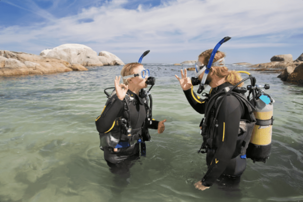 Two scuba divers in wetsuits stand in shallow, clear water near a rocky shore, facing each other and making the “OK” hand signal, with blue skies and large rocks in the background.