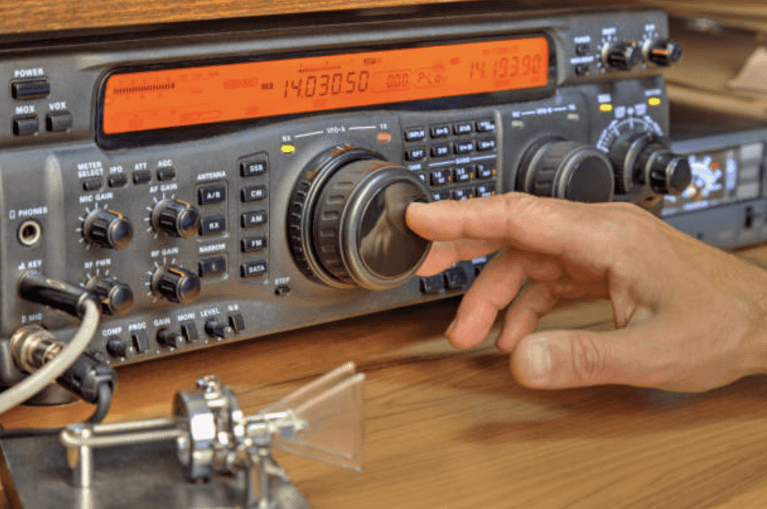 A hand adjusts the main tuning dial on a ham radio transceiver sitting on a wooden desk, with various buttons, knobs, and an orange digital display visible on the radio. A Morse code keyer is also seen in the foreground.