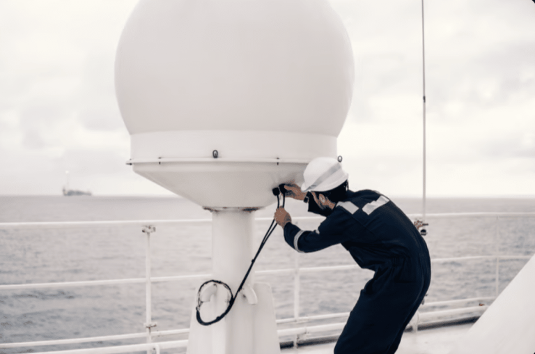 A person in a navy coverall and white hard hat works on a large white satellite dome on a ship's deck, with the ocean and an offshore platform visible in the background.