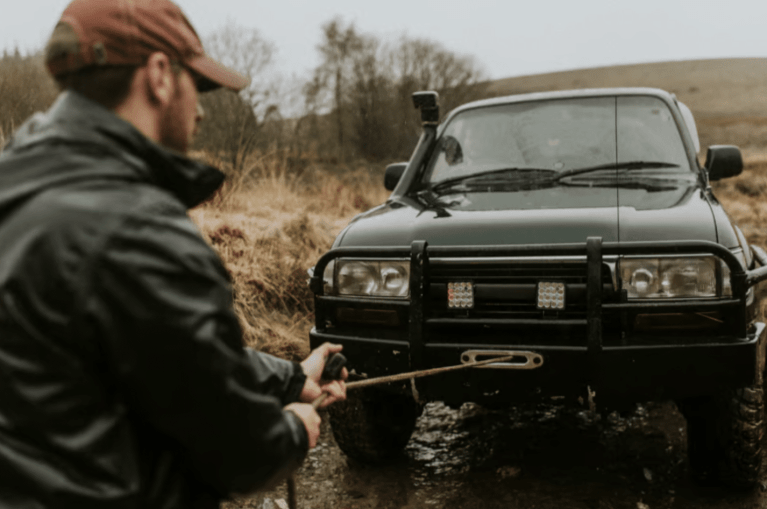 A person in a maroon cap and dark jacket prepares to attach a tow strap to the front of a black off-road vehicle on a muddy track in a rural, grassy area.