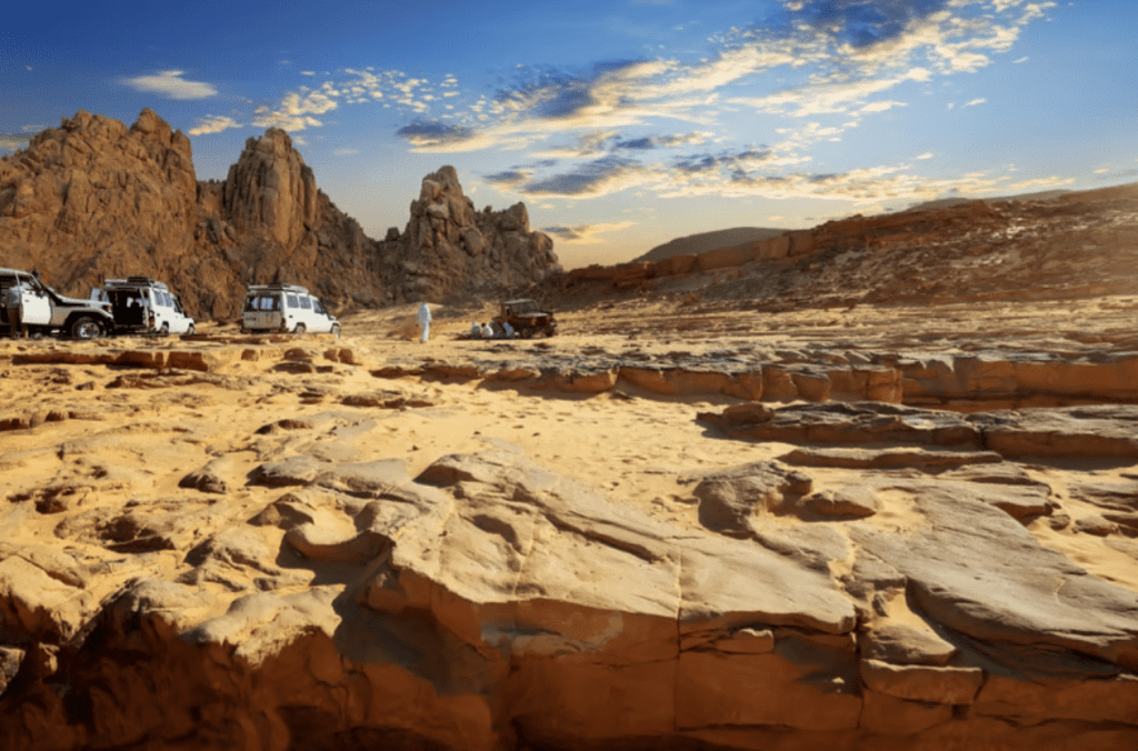 Several off-road vehicles are parked on a rocky, desert landscape with rugged cliffs in the background under a partly cloudy blue sky. The scene is bathed in warm sunlight.