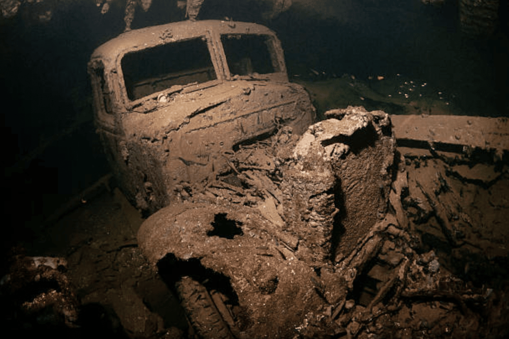 Screenshot 2026-03-31 at 214906 - N9BO℠ | Global Underwater Services Ltd An old, rusted lorry covered in sediment lies underwater, partially buried on the seabed with decaying, broken parts visible in the dim light.