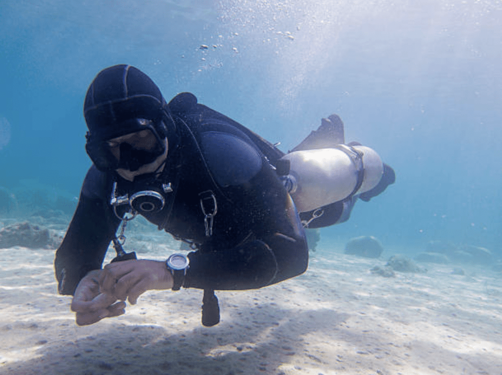 Screenshot 2026-03-31 at 222117 - N9BO℠ | Global Underwater Services Ltd A scuba diver in a black wetsuit and diving gear swims just above the sandy sea floor, surrounded by clear blue water and some rocks in the background.