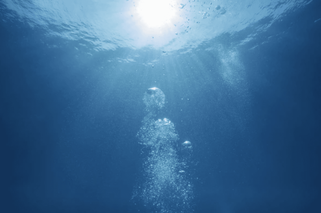Screenshot 2026-03-31 at 224725 - N9BO℠ | Global Underwater Services Ltd Sunlight filters through the surface of clear blue water as air bubbles rise towards the top, creating a serene underwater scene.