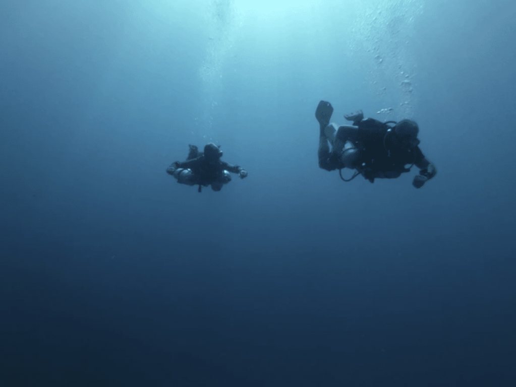 Screenshot 2026-03-31 at 224814 - N9BO℠ | Global Underwater Services Ltd Two scuba divers swimming underwater in deep blue ocean, with light filtering down from the surface above them.
