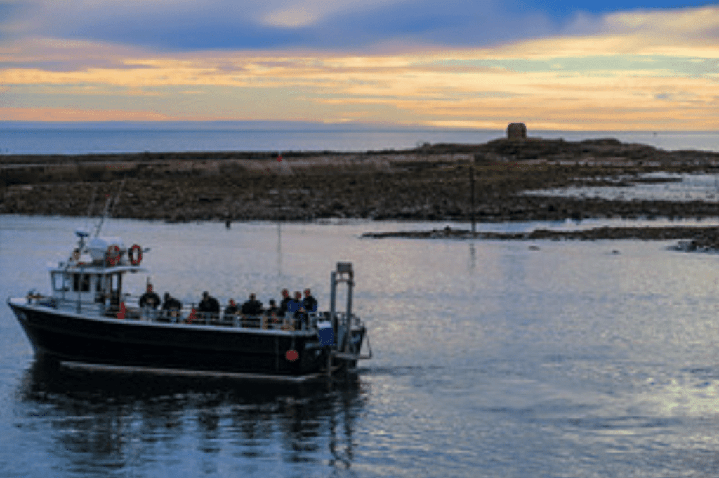 A small boat carrying people sails on calm water near a rocky shore at sunset, with a colourful sky and an island with a stone structure in the background.
