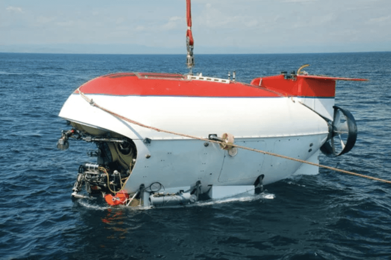 A red and white submersible is partially out of the water, suspended by a crane and cables, with the sea and a distant shoreline visible in the background.