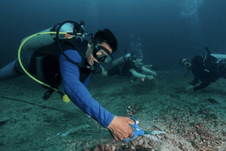 A scuba diver uses scissors to cut a fishing net on the seabed while two other divers assist in the background, surrounded by underwater rocks and marine debris.