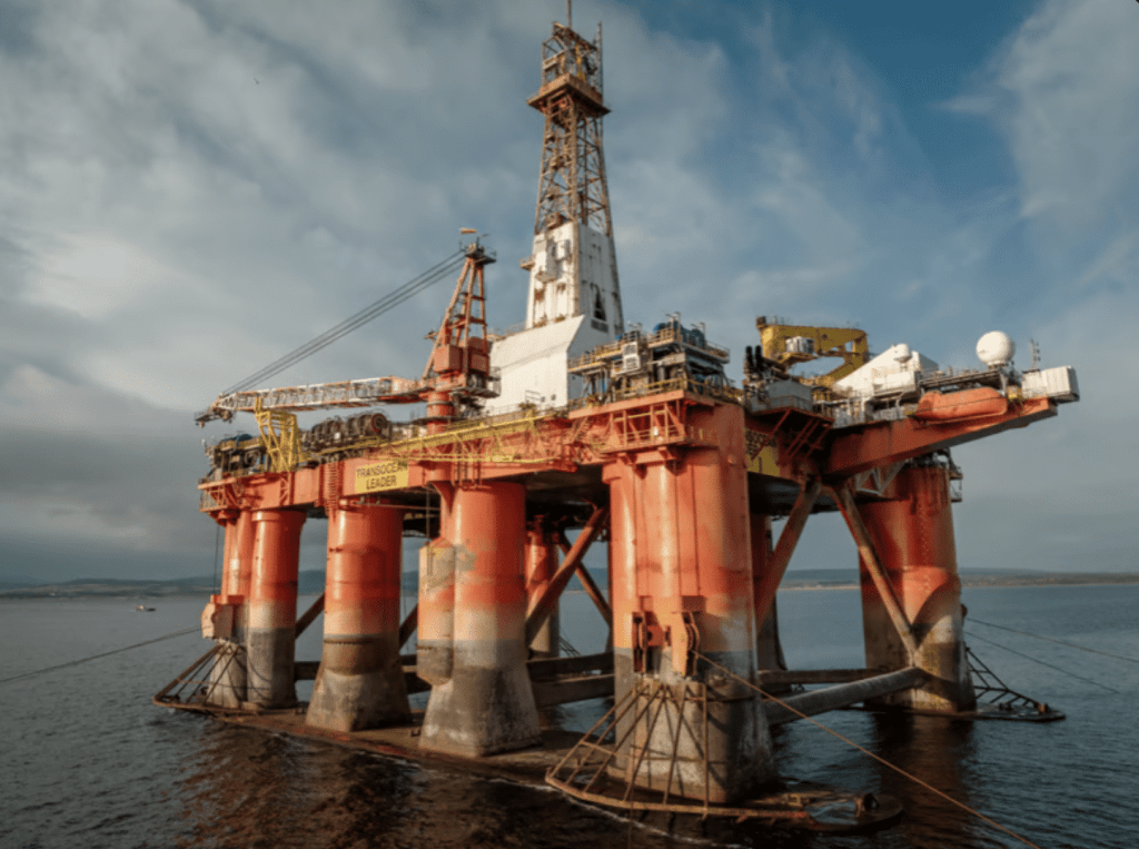 Screenshot 2026-03-31 at 235520 - N9BO℠ | Global Underwater Services Ltd An offshore oil rig with red and yellow structure stands anchored in the sea under a partly cloudy sky, with cables stretching to the water and land faintly visible in the background.