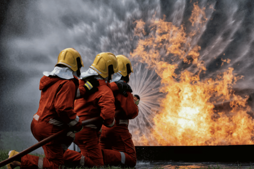 Screenshot 2026-03-31 at 235554 - N9BO℠ | Global Underwater Services Ltd Three firefighters in orange suits and yellow helmets kneel on grass, aiming a hose that sprays water at a large, intense fire with billowing smoke.