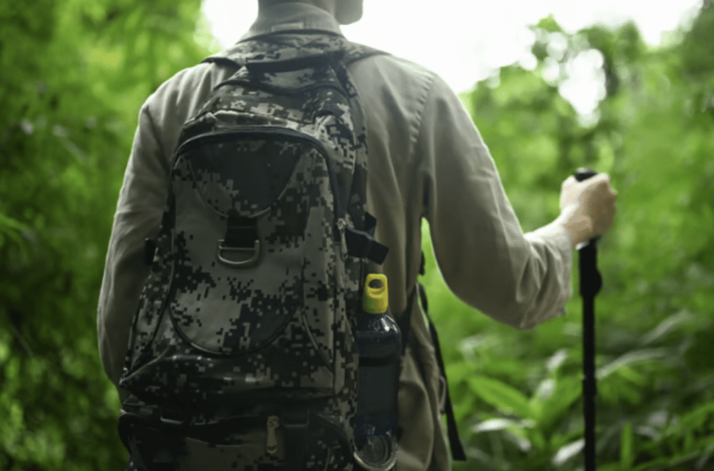 A person with a camouflage rucksack and walking stick hikes through lush green foliage, seen from behind in a forest or jungle setting.