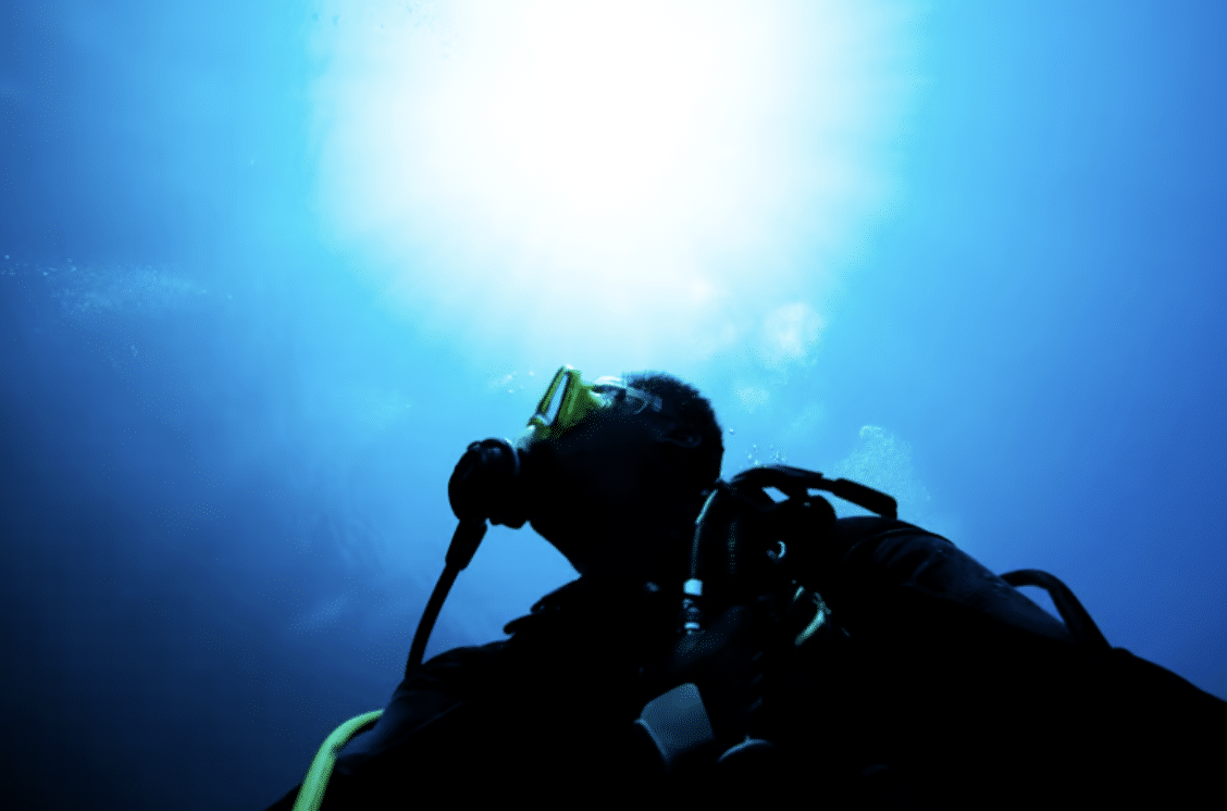 Screenshot 2026-04-01 at 002222 - N9BO℠ | Global Underwater Services Ltd A scuba diver underwater is silhouetted against bright sunlight filtering through the blue water above, wearing a mask and breathing apparatus.