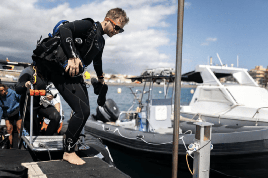 Screenshot 2026-04-01 at 002242 - N9BO℠ | Global Underwater Services Ltd A person in a wetsuit and diving gear steps barefoot off a jetty towards a black inflatable boat on the water, with another person and several boats in the background under a partly cloudy sky.