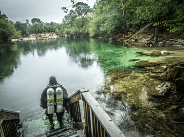 A scuba diver wearing two air cylinders walks down wooden steps into a clear, calm river surrounded by lush green trees and rocky shoreline.