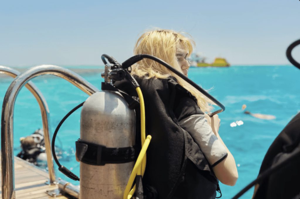 A person with blonde hair wearing scuba gear sits at the edge of a boat, looking towards the bright turquoise sea under a clear sky, with other divers and a distant boat visible in the background.