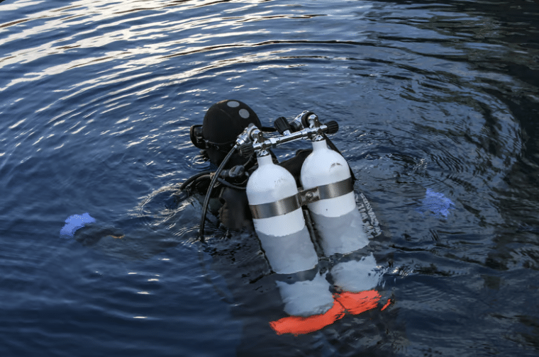 Screenshot 2026-04-01 at 003616 - N9BO℠ | Global Underwater Services Ltd A scuba diver wearing a black wetsuit and twin air cylinders is partly submerged in calm, dark blue water, creating circular ripples around them. The diver’s gloves and a hint of orange kit are visible.