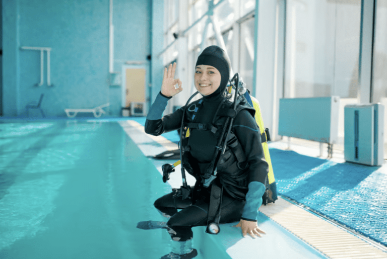 A person in a wetsuit and scuba gear sits on the edge of an indoor swimming pool, smiling and making an OK hand gesture, ready for diving practice. Bright daylight fills the spacious pool area.