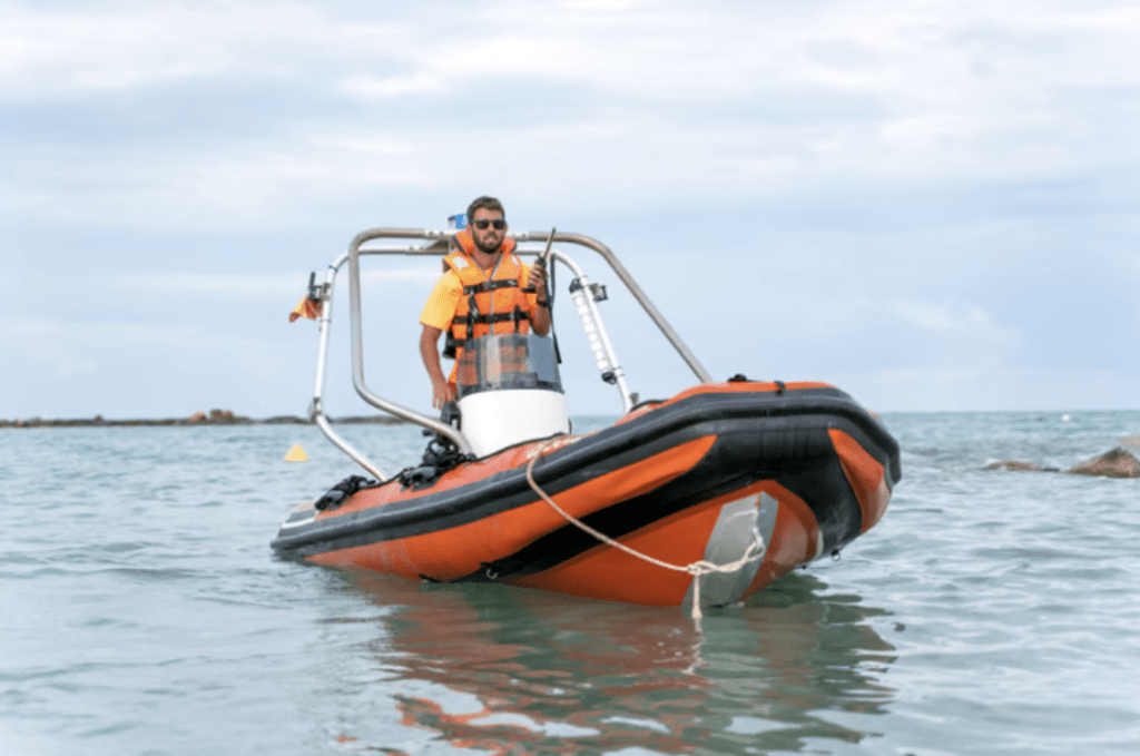 Screenshot 2026-04-01 at 004950 - N9BO℠ | Global Underwater Services Ltd A man wearing a life jacket stands on an orange inflatable boat floating on calm water near the shore, under a cloudy sky. The boat is equipped with instruments and has a rope tied to its front.