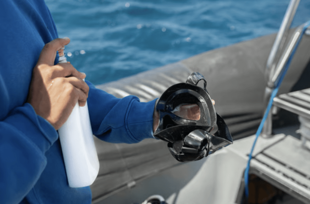 Screenshot 2026-04-01 at 005001 - N9BO℠ | Global Underwater Services Ltd A person wearing a blue jumper holds a black snorkelling mask in one hand and a white spray bottle in the other, standing on a boat with blue sea water in the background.