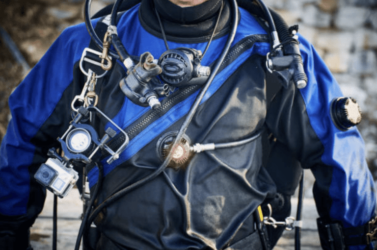 A person wearing a blue and black drysuit with scuba diving equipment, including a regulator, pressure gauge, and an underwater camera attached to their chest. The background is blurred.