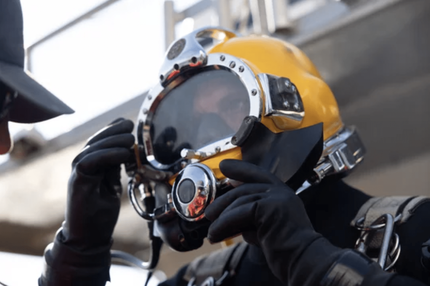 Screenshot 2026-04-01 at 020605 - N9BO℠ | Global Underwater Services Ltd A person wearing a yellow commercial diving helmet and black gloves adjusts their mask, preparing for an underwater operation. Another figure in a hat is partially visible on the left.