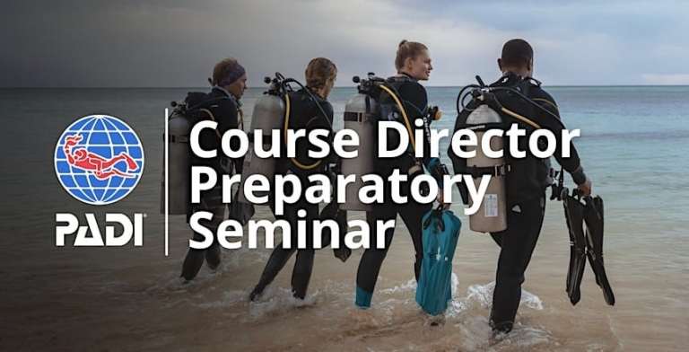 Four scuba divers in wetsuits carry cylinders and flippers as they walk towards the sea. The PADI logo and text read “Course Director Preparatory Seminar.” The sky is cloudy, and the shoreline is visible.