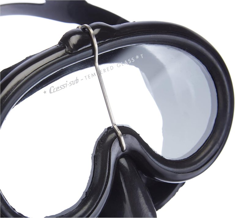 Close-up of a black diving mask with toughened glass lenses. The mask has a metal wire clip across the nose bridge area, and the words Cressi-sub TOUGHENED GLASS are visible on one of the lenses.