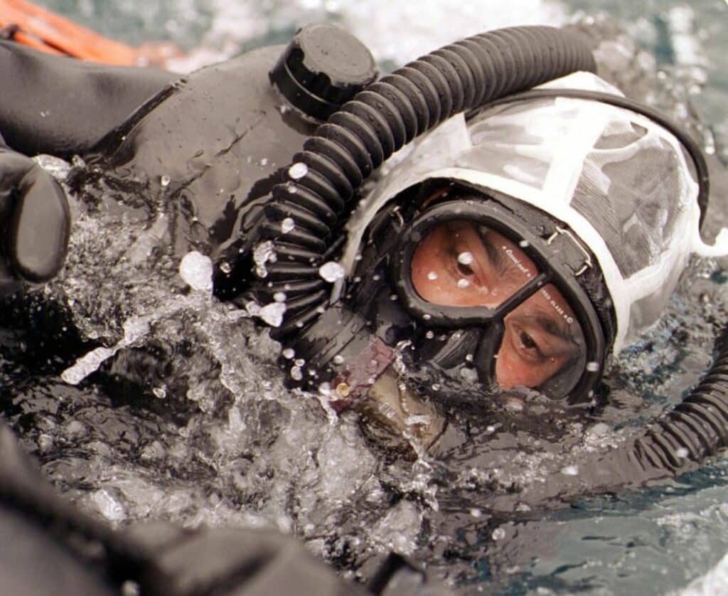 A diver wearing a full-face mask and breathing apparatus is partially submerged in water, with bubbles and splashes around them. The diver's face is visible through the goggles.