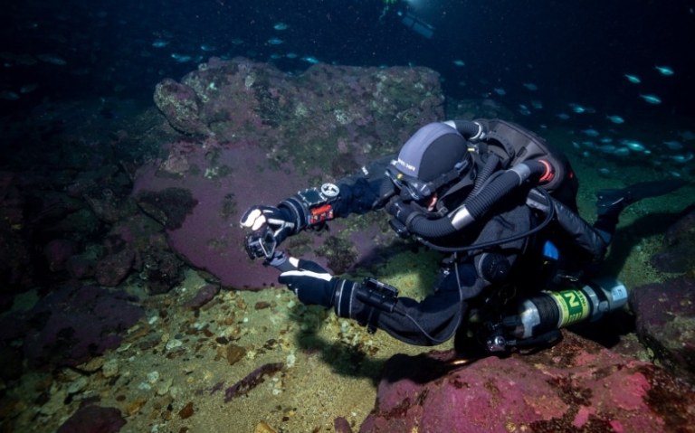 A scuba diver wearing a full-face mask and technical diving gear explores underwater rocks, holding a camera and pointing at something on the sea floor. Small fish swim in the background.