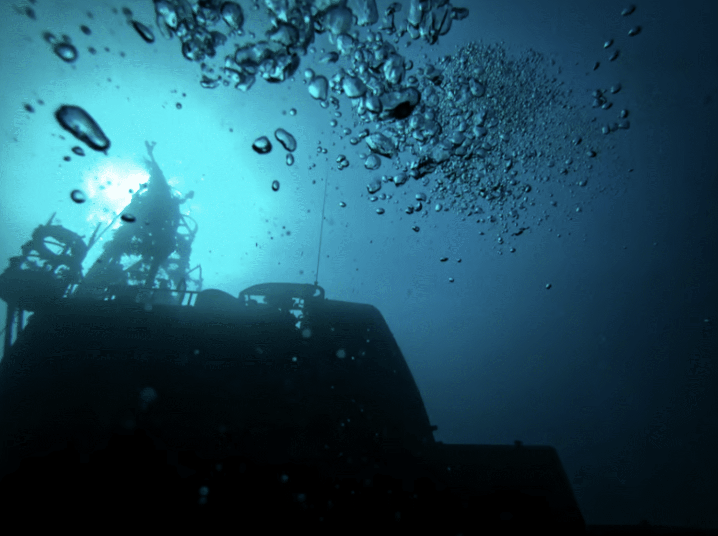 Screenshot 2026-04-01 at 091837 - N9BO℠ | Global Underwater Services Ltd Underwater view of a shipwreck with sunlight filtering from above, bubbles rising towards the surface, and the silhouette of the vessel partially visible in the deep blue water.