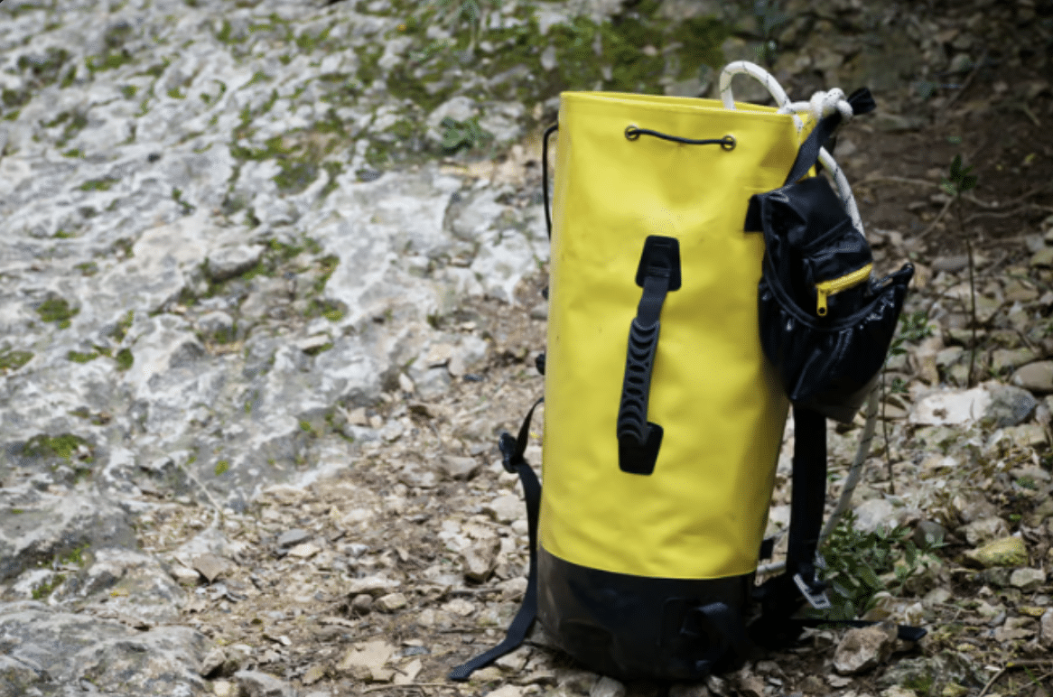 Screenshot 2026-04-01 at 093512 - N9BO℠ | Global Underwater Services Ltd A bright yellow waterproof rucksack with black straps and a side pocket sits on rocky, uneven ground outdoors, surrounded by small stones and patches of grass.