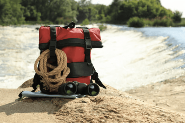 A red rucksack with black straps, a coiled rope, a pair of binoculars, and a map rests on a rock by a river with fast-flowing water and green trees in the background.