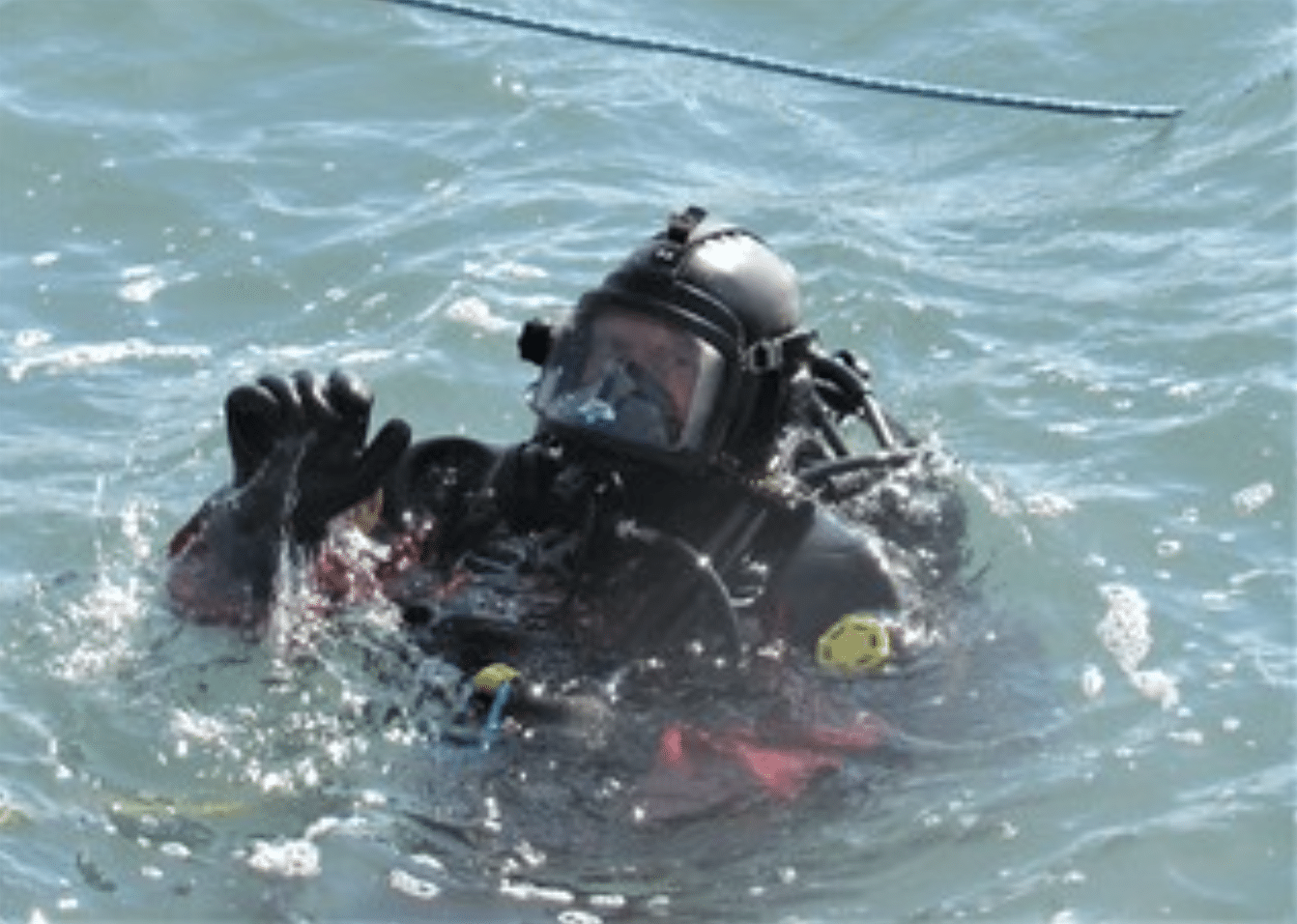 A diver wearing a full-face mask and black wetsuit emerges from blue water, raising one hand in a gesture. A blue rope floats on the water's surface nearby.