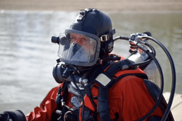 A scuba diver wearing a full-face mask, helmet, and bright red drysuit is preparing to enter the water, with scuba cylinders visible on their back and a calm shoreline in the background.