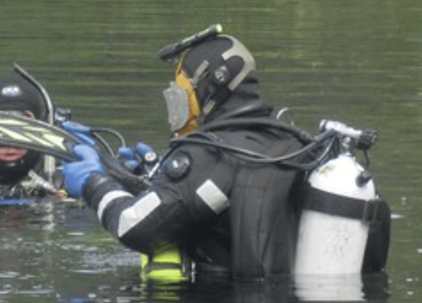 A scuba diver in a black wetsuit and blue gloves is standing in water, wearing a mask and an oxygen cylinder, and interacting with another diver.