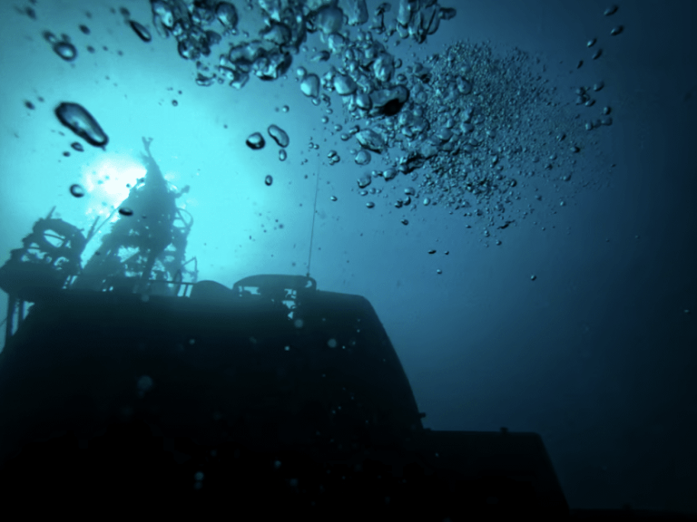 Bubbles rise towards the surface above the dark silhouette of a submerged structure, possibly a shipwreck, illuminated by faint blue underwater light.