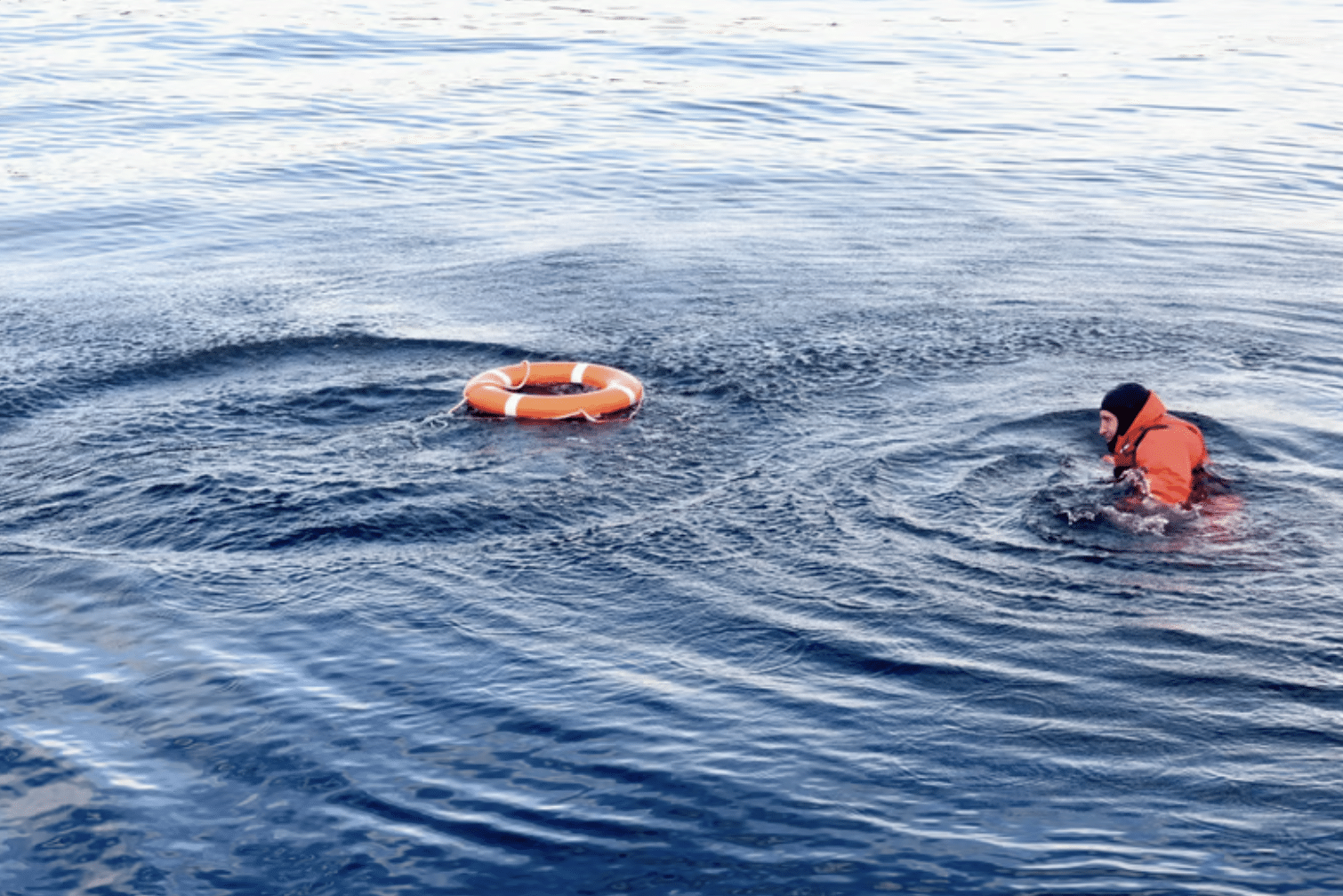 Screenshot 2026-04-01 at 104237 - N9BO℠ | Global Underwater Services Ltd A person wearing an orange lifejacket is swimming in open water towards an orange lifebuoy floating nearby. The water is calm with gentle ripples.