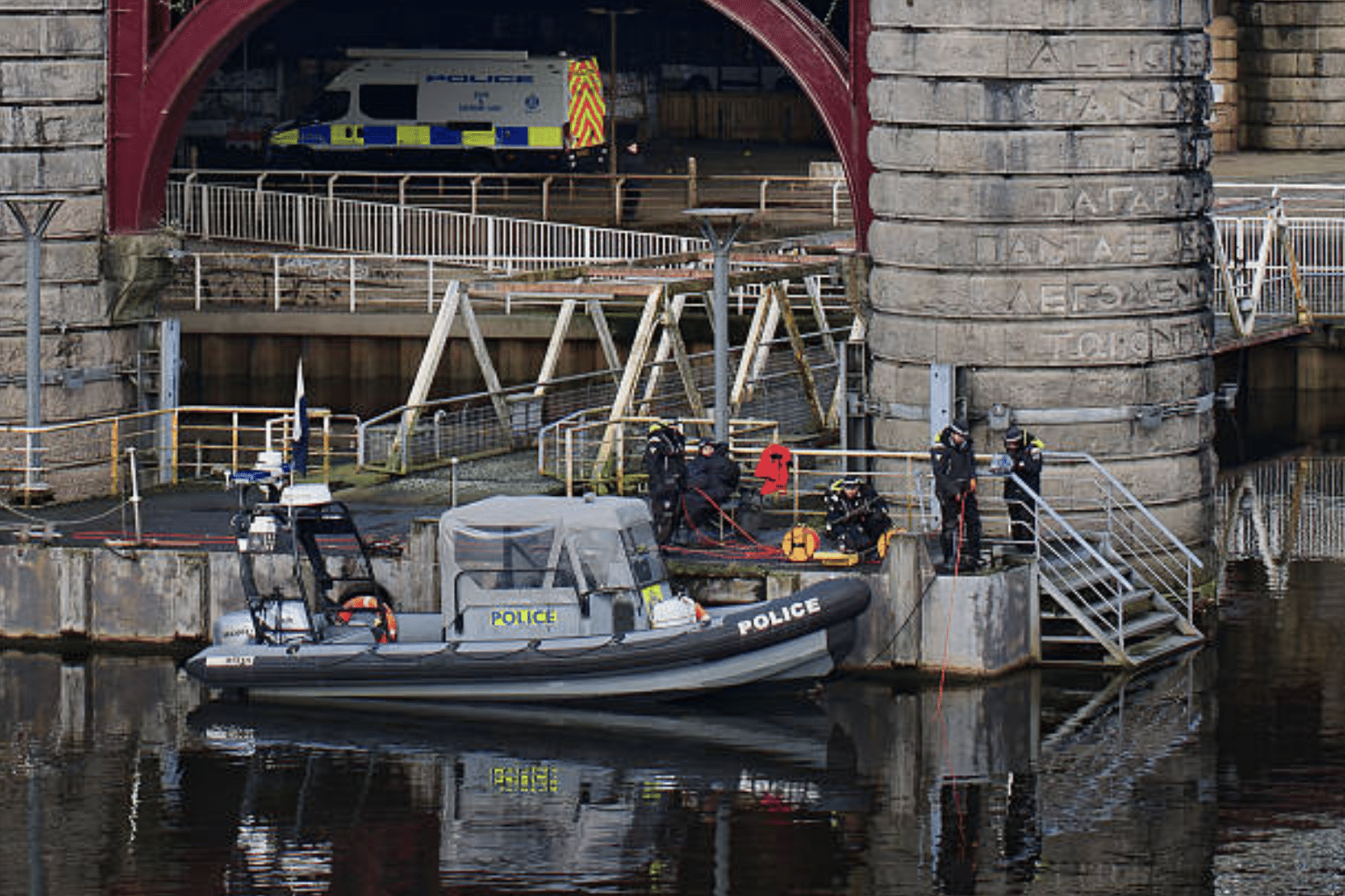 Screenshot 2026-04-01 at 104410 - N9BO℠ | Global Underwater Services Ltd A police boat is moored beside a stone bridge pier over a river, with several officers and divers in gear standing on nearby steps and a police van parked under the bridge.