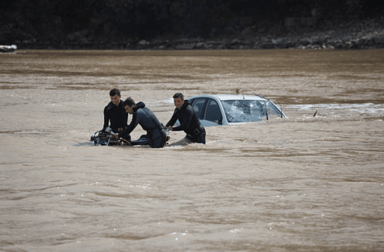 Three rescue workers stand in knee-deep brown floodwater, operating equipment near a partially submerged car, with only the top of the car visible above the water. Trees and a rocky shoreline are in the background.