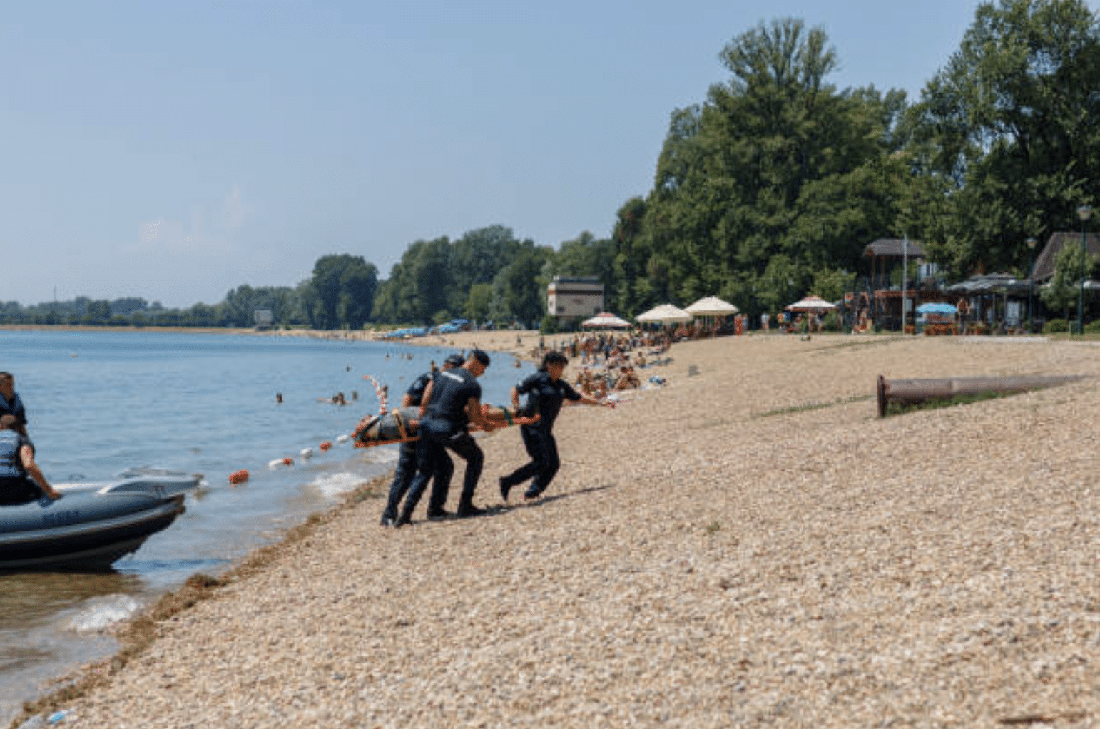 Screenshot 2026-04-01 at 105823 - N9BO℠ | Global Underwater Services Ltd Three emergency responders carry a person on a stretcher along a pebbled beach, with people and parasols in the background and a boat by the shore on a sunny day.