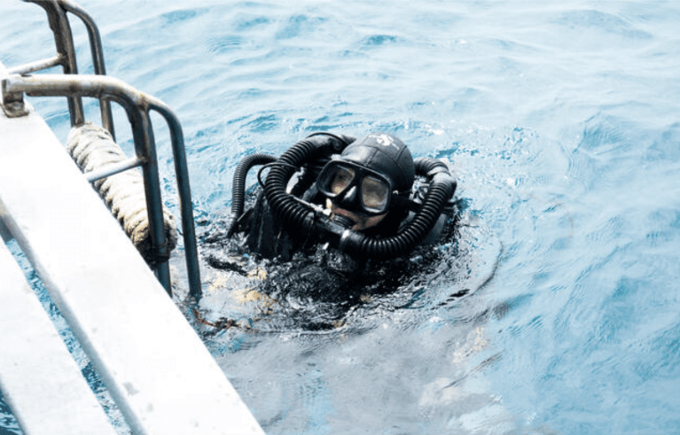 A scuba diver wearing a full-face mask and rebreather gear floats in blue water near a metal ladder attached to a boat.