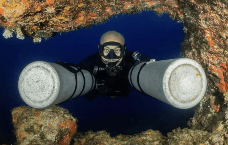 A scuba diver with two large oxygen cylinders swims through a rocky, coral-covered underwater arch, looking directly at the camera against a deep blue backdrop.